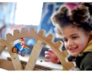 LEGO City 60298 Rocket Stunt Bike minifigure on blue motorcycle balancing on wooden gear structure, smiling child.