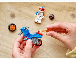 Hands assembling a blue LEGO Rocket Stunt Bike. Minifigure, extra wheel, and hairpiece on wooden background.