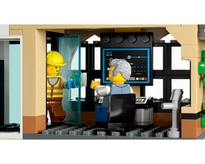 Two LEGO minifigures in a train station: a construction worker entering an office, and an older man at a desk with a monitor.