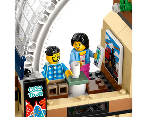 LEGO minifigures in a train station cafe. A man sits with coffee, a woman holds a smartphone near a train poster and city tour sign.