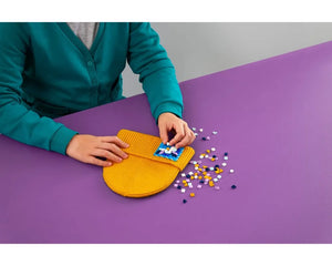 Hands decorating a yellow knitted LEGO Dots stitch-on patch with colorful tiles on a purple table.