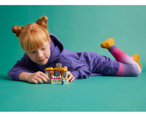 A child with red hair plays with the LEGO Friends 42608 Tiny Accessories Store set while lying on a teal background.