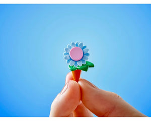 A hand holds a small light blue LEGO flower with a pink center, green leaf, and orange stem against a bright blue sky.