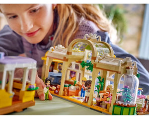 Girl interacting with LEGO Friends 42671 Plant Café & Flower Shop set, featuring a detailed building, minifigures, plants, and a tram.