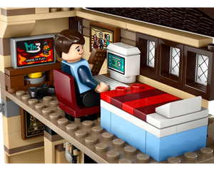 A LEGO minifigure sits at a desk with a monitor displaying a game, next to a bed in a room with windows.