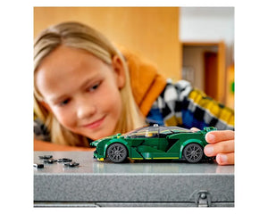 Young girl looks intently at a green LEGO Speed Champions Lotus Evija car, holding its rear. Small LEGO pieces are on a grey surface.