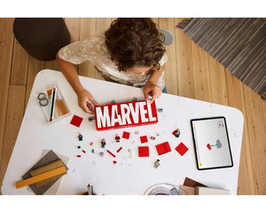 Overhead view of a child building a red and white LEGO Marvel logo. Loose bricks, minifigures, and a tablet on a white table.