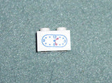 White LEGO 1x2 brick with a clock sticker pattern. Blue numbers 3, 6, 9, 12, and red hour and minute hands.