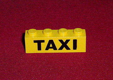 Yellow LEGO 1x4 brick with a black 'TAXI' pattern printed on its side, resting on a reddish-brown background.