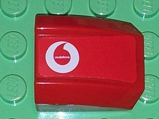 A red LEGO curved 2x2 slope brick with a white Vodafone logo sticker, displayed on a green studded base.