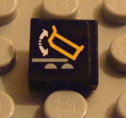 Black LEGO 1x1 tile 3070pb042 with a sticker featuring a yellow dump truck and white arrows indicating a tilting/dumping action.