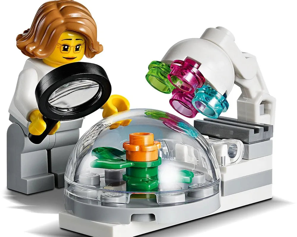 LEGO minifigure scientist with magnifying glass studies a plant in a dome next to a microscope.