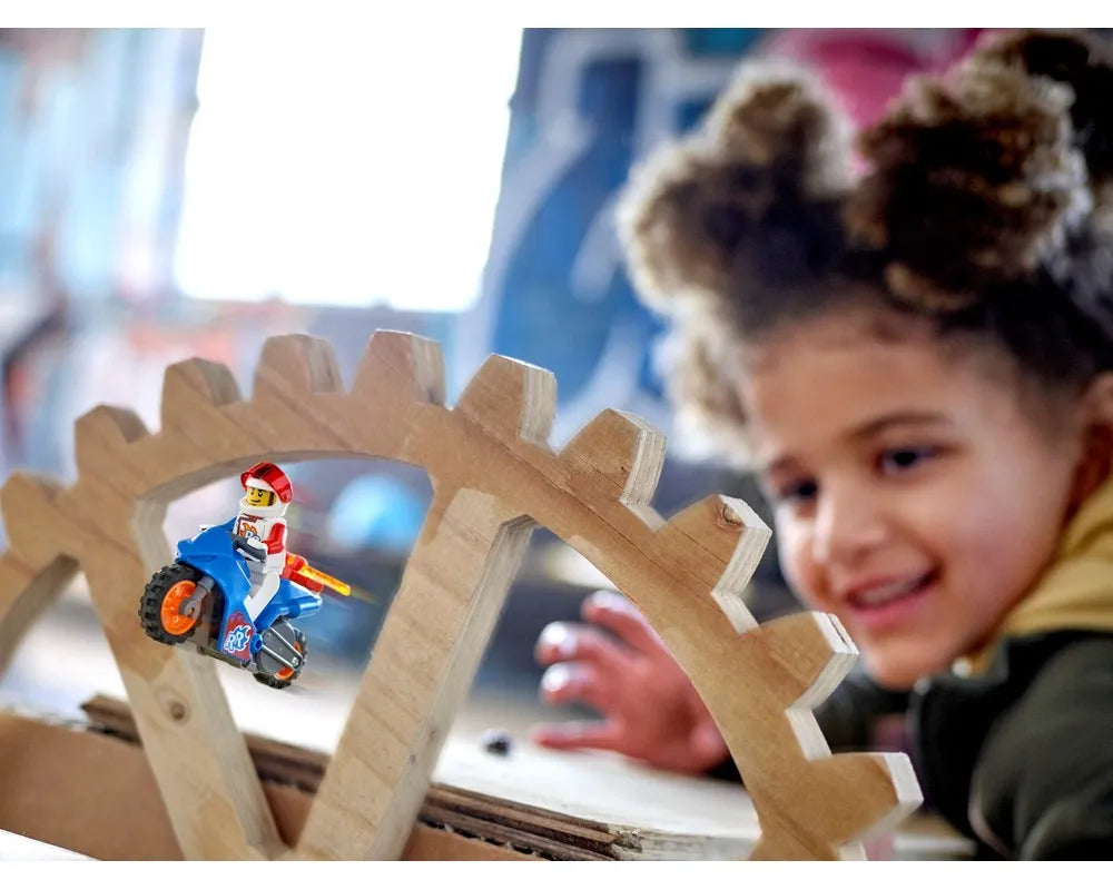 LEGO City 60298 Rocket Stunt Bike minifigure on blue motorcycle balancing on wooden gear structure, smiling child.