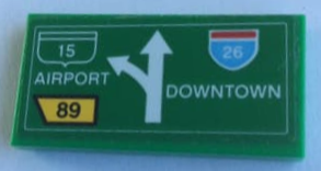 Green LEGO 2x4 tile with road sign pattern: white split arrow, text 'AIRPORT', 'DOWNTOWN', highway shields '15', '26', and route '89'.