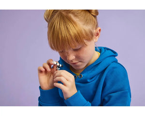 A young girl with red hair and freckles in a blue hoodie intently looks at a small LEGO telescope on a white stand.