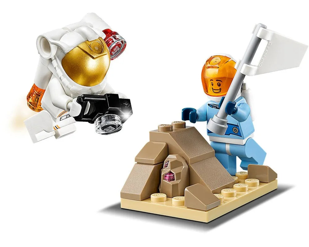Two LEGO astronauts: one in white taking a photo, one in blue planting a flag next to a rock with a purple crystal.