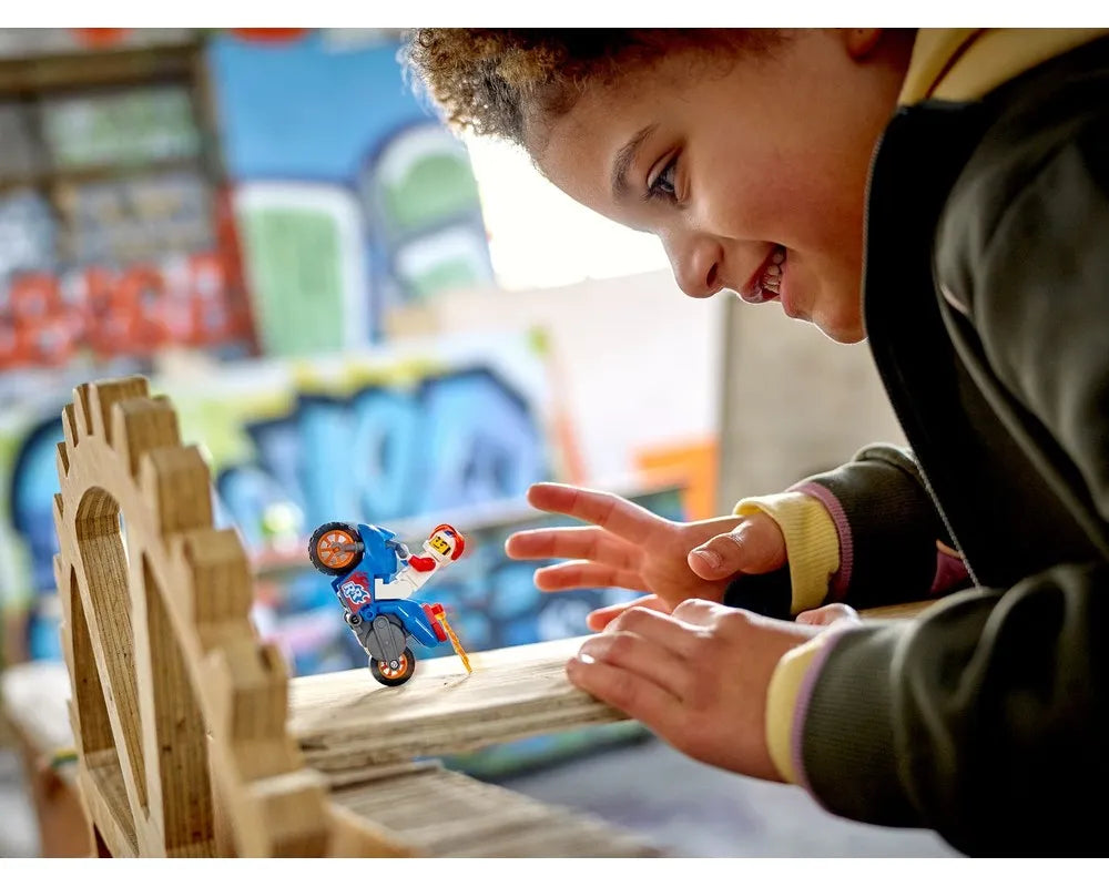 Smiling child plays with LEGO City 60298 Rocket Stunt Bike, a blue motorcycle with minifigure on a wooden ramp.