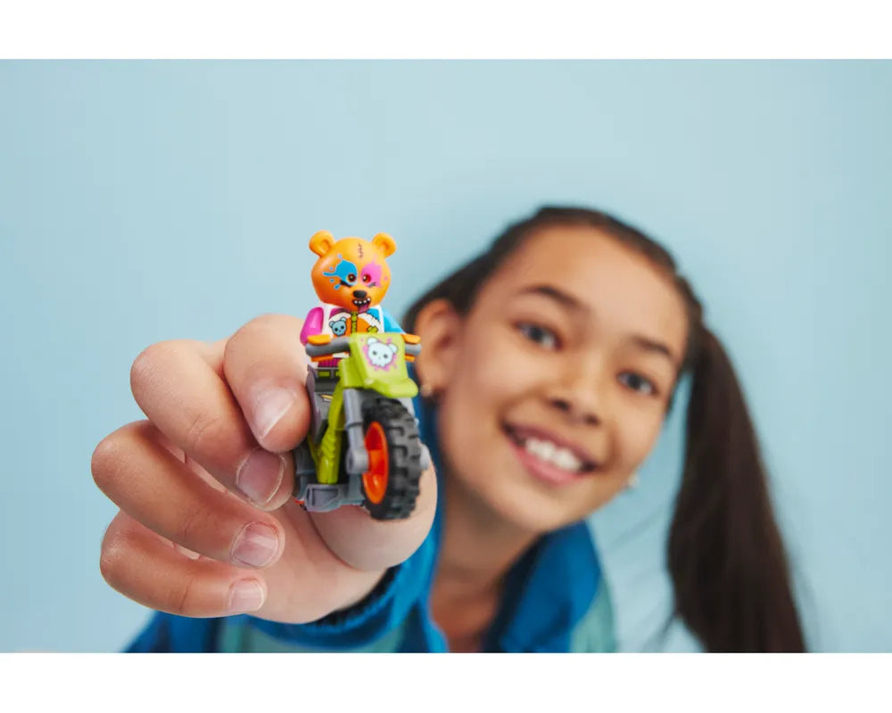 A smiling girl holds a LEGO City 60356 Bear Stunt Bike, featuring an orange bear minifigure on a green bike.
