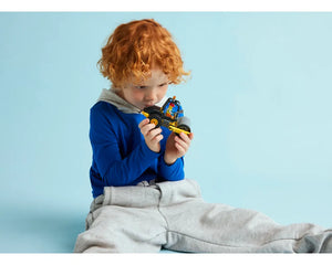 A red-haired child in blue and grey clothes sits on a blue floor, looking intently at a black and yellow LEGO City 60401 Construction Steamroller.