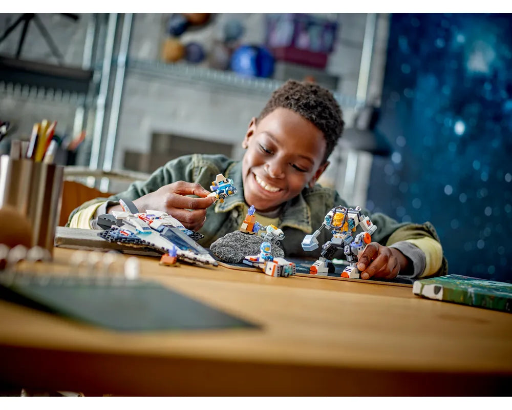 A smiling boy plays with LEGO City space sets, including a spaceship, a mech, and a small vehicle, on a wooden desk.