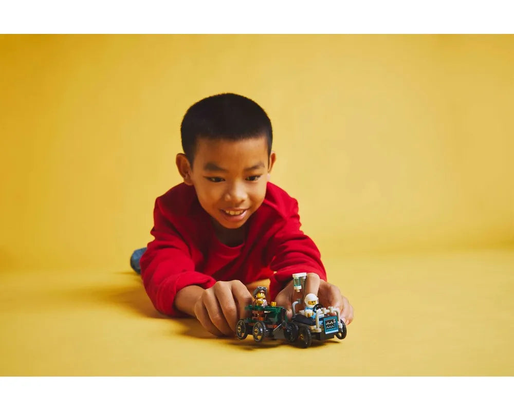 A smiling boy in a red shirt plays with two small LEGO vehicles with minifigures on a yellow floor.