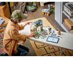 Child assembling LEGO City 60469 Central Train Station set on a table with a tablet.