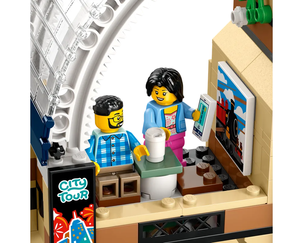 LEGO minifigures in a train station cafe. A man sits with coffee, a woman holds a smartphone near a train poster and city tour sign.