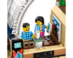 LEGO minifigures in a train station cafe. A man sits with coffee, a woman holds a smartphone near a train poster and city tour sign.