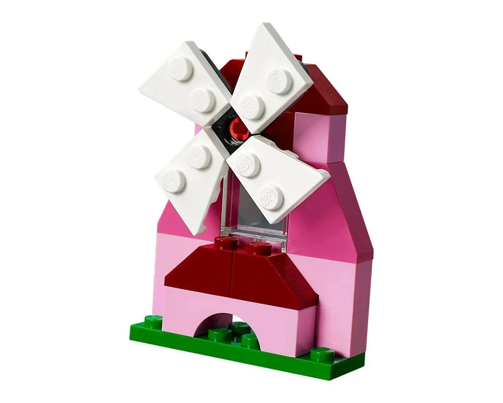 A small LEGO windmill, built from pink and dark red bricks, with four white rotating blades, standing on a green baseplate.