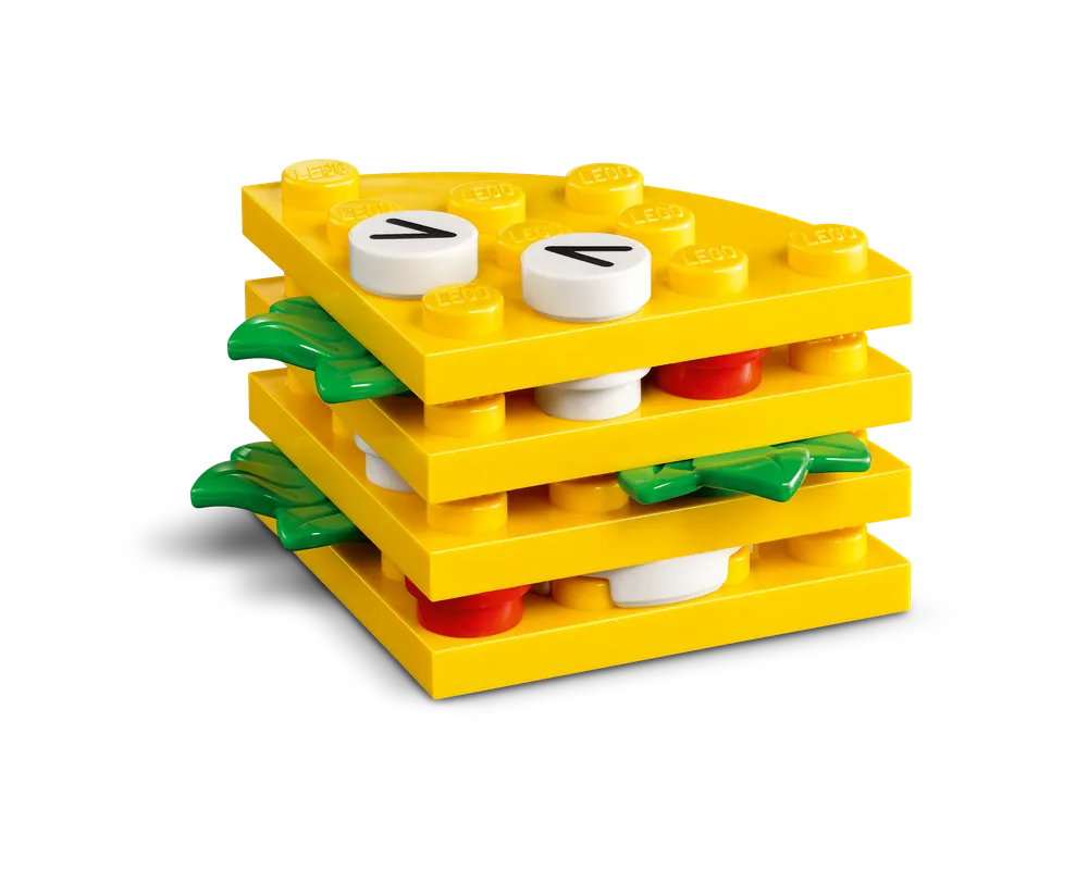 A layered LEGO food build featuring yellow plates, green leaf elements, red round bricks, and white round bricks with black markings.