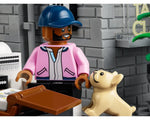 A LEGO minifigure with a pink jacket and blue cap stands with a tan French bulldog. Grey brick walls and a brown desk are visible.