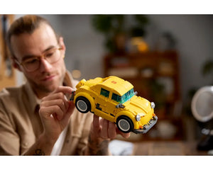 Man in glasses holding a yellow LEGO Creator 10338 Bumblebee car, adding a piece to the back.