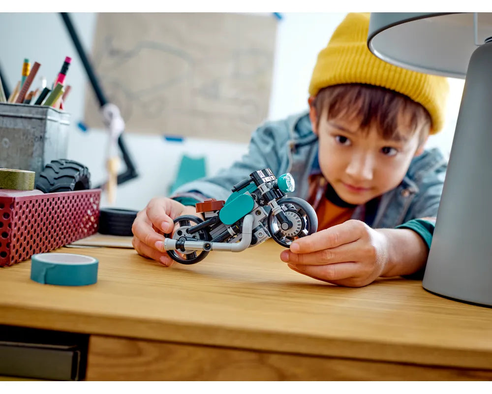 Boy in yellow beanie holds a teal LEGO Creator 31135 Vintage Motorcycle on a wooden desk with craft supplies.