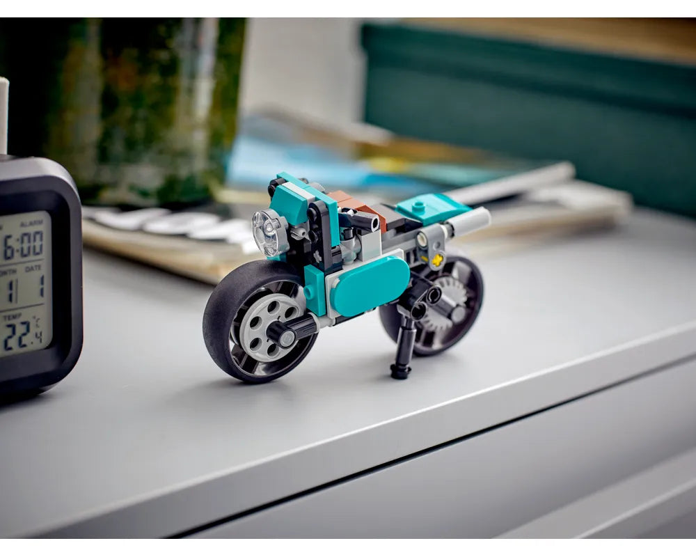 A teal and grey LEGO Creator 31135 Vintage Motorcycle on a light grey surface next to a digital clock.