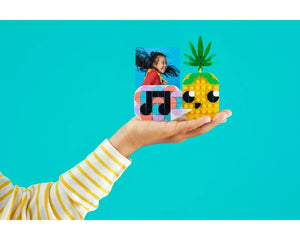 A hand holds a LEGO DOTS photo holder with a child's picture and a yellow pineapple character, on a teal background.