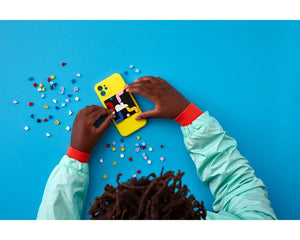 Hands customizing a yellow phone case with colorful LEGO DOTS tiles from the 41954 set on a blue background.