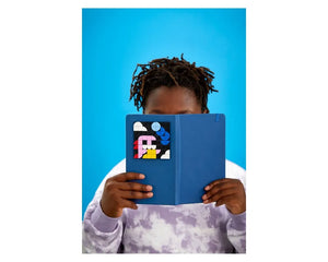 Person holding a blue notebook featuring a LEGO Dots 41954 adhesive patch with colorful abstract design.