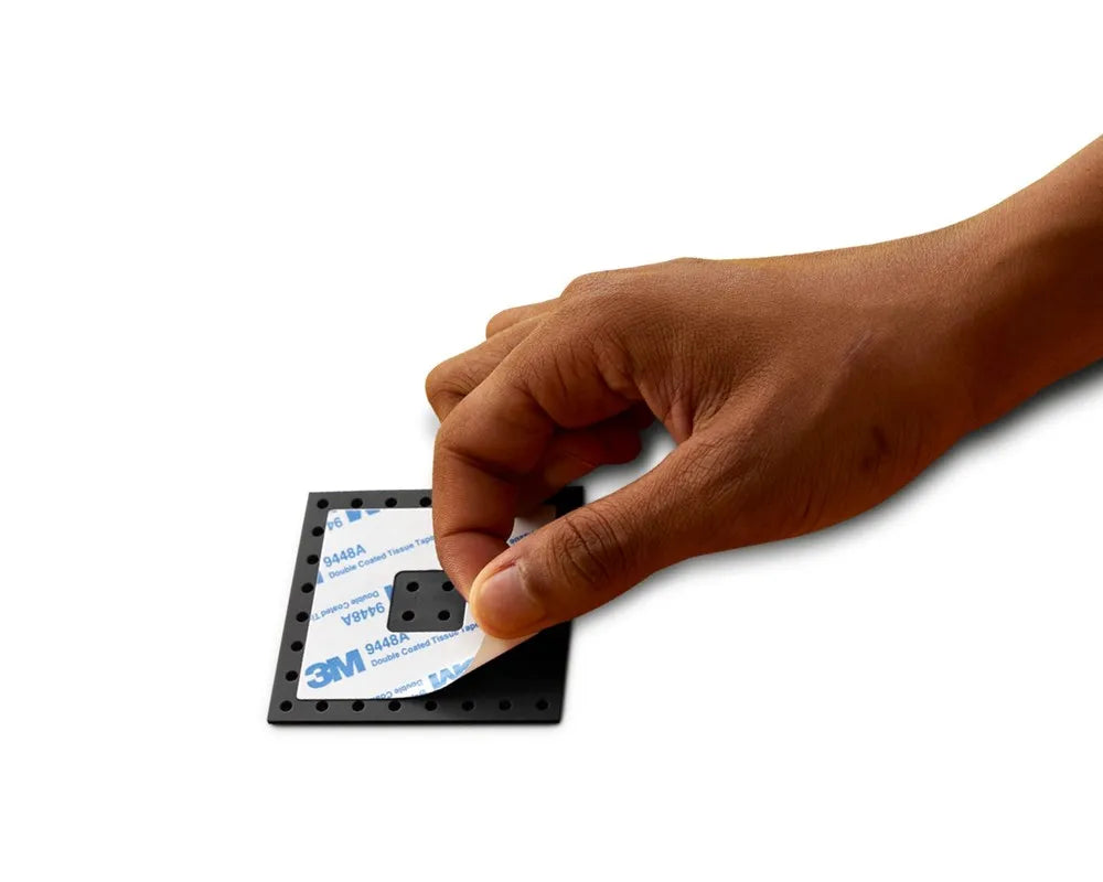 Hand peeling backing off a black square LEGO Dots adhesive patch with multiple holes, revealing 3M double-sided tape.