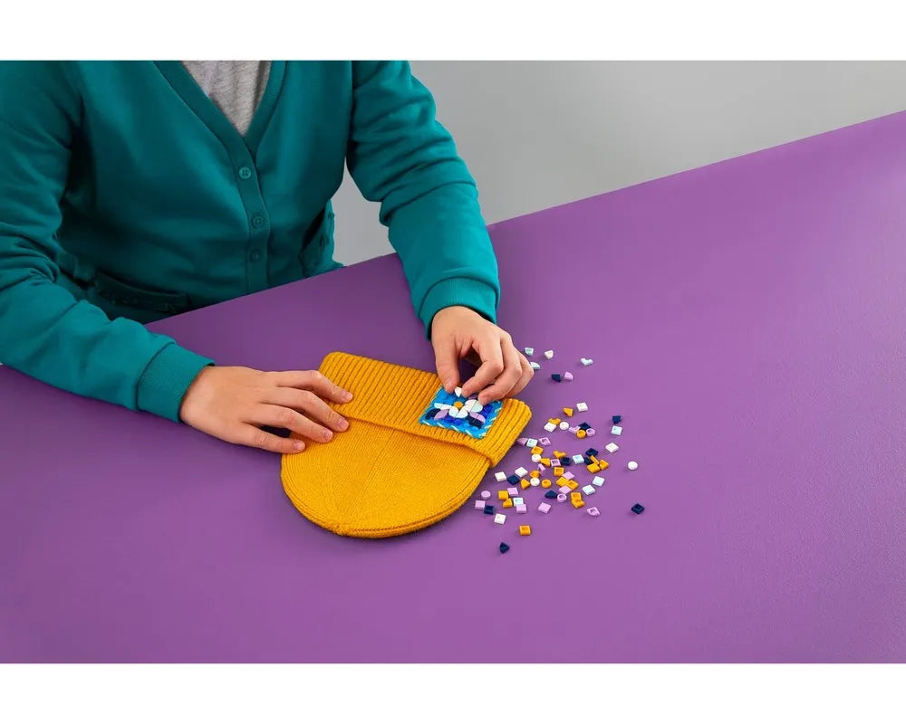 Hands decorating a yellow knitted LEGO Dots stitch-on patch with colorful tiles on a purple table.