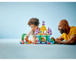 A young child and an adult man play with the colorful LEGO DUPLO Ariel's Magical Underwater Palace set, a blue, yellow, and purple castle.