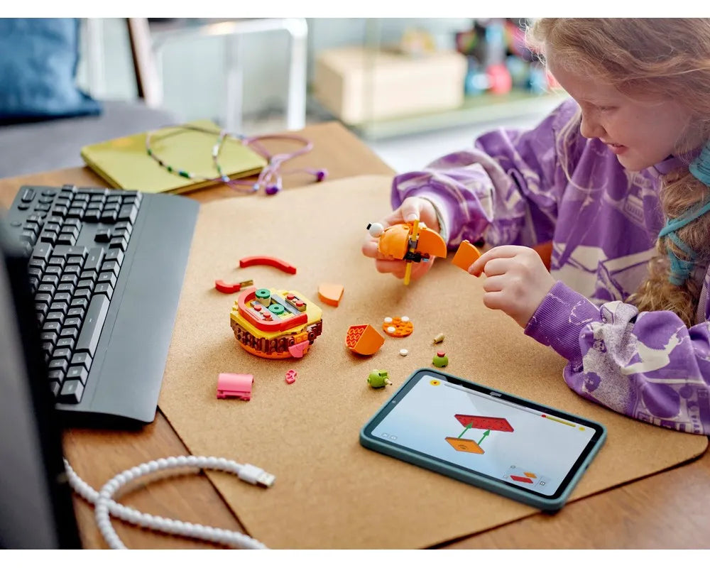 Girl assembling LEGO Durrr Burger set, an orange creature, and other bricks. Tablet shows build steps.