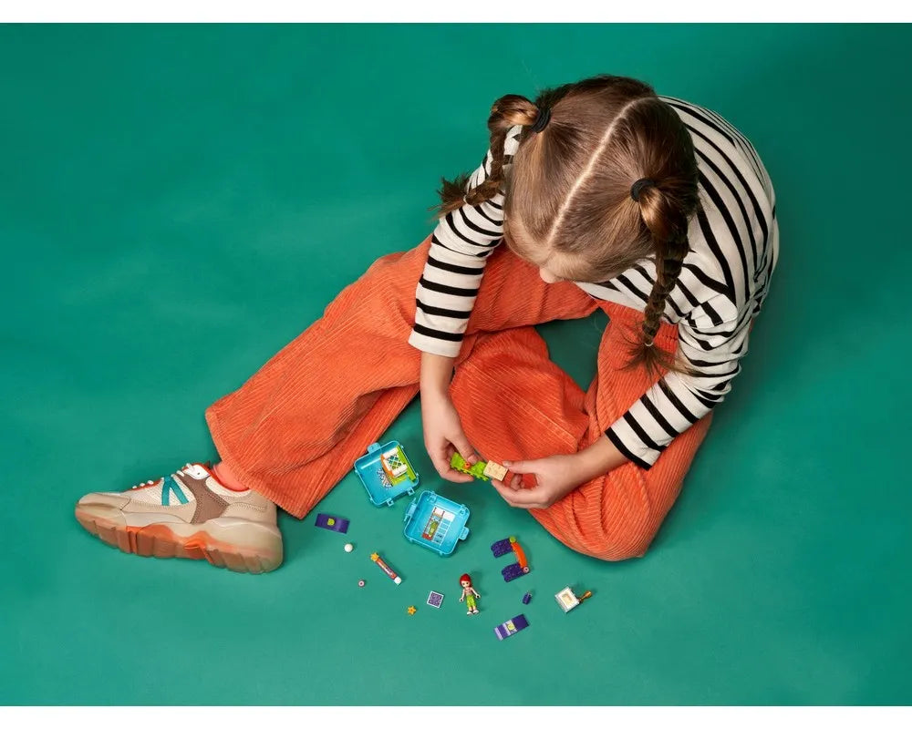 Child sits on green floor, playing with LEGO Friends Mia's Soccer Cube, holding green bricks. Other pieces and minifigure are spread.