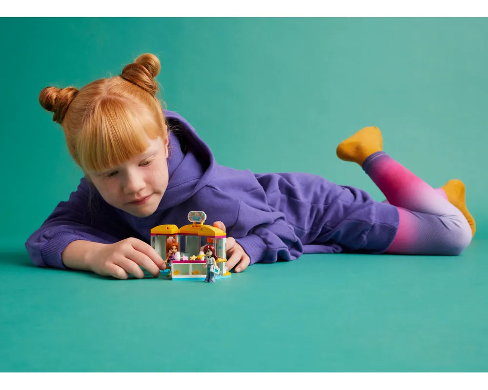 A child with red hair plays with the LEGO Friends 42608 Tiny Accessories Store set while lying on a teal background.