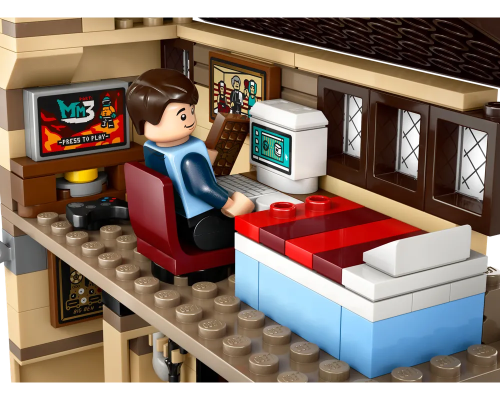 A LEGO minifigure sits at a desk with a monitor displaying a game, next to a bed in a room with windows.