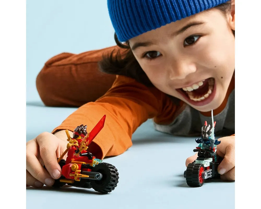 A smiling child plays on the floor with two LEGO Ninjago motorcycles. One is red with Kai, the other dark colored.