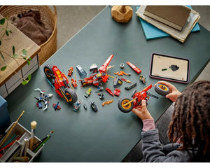 A child building a red LEGO Ninjago 71844 Ninja Combat Vehicle with digital instructions on a tablet, surrounded by minifigures and another large red LEGO vehicle.