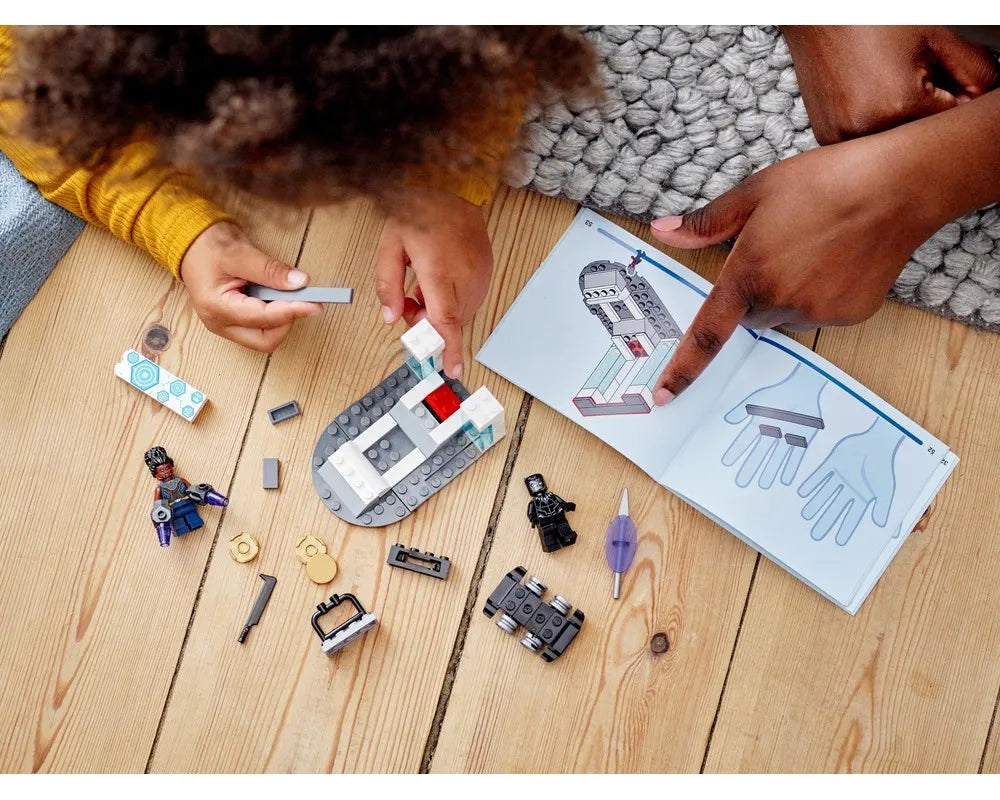 A child and an adult building LEGO Super Heroes 76212 Shuri's Lab on a wooden floor, with minifigures and instructions.