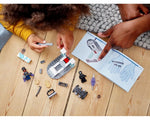 A child and an adult building LEGO Super Heroes 76212 Shuri's Lab on a wooden floor, with minifigures and instructions.