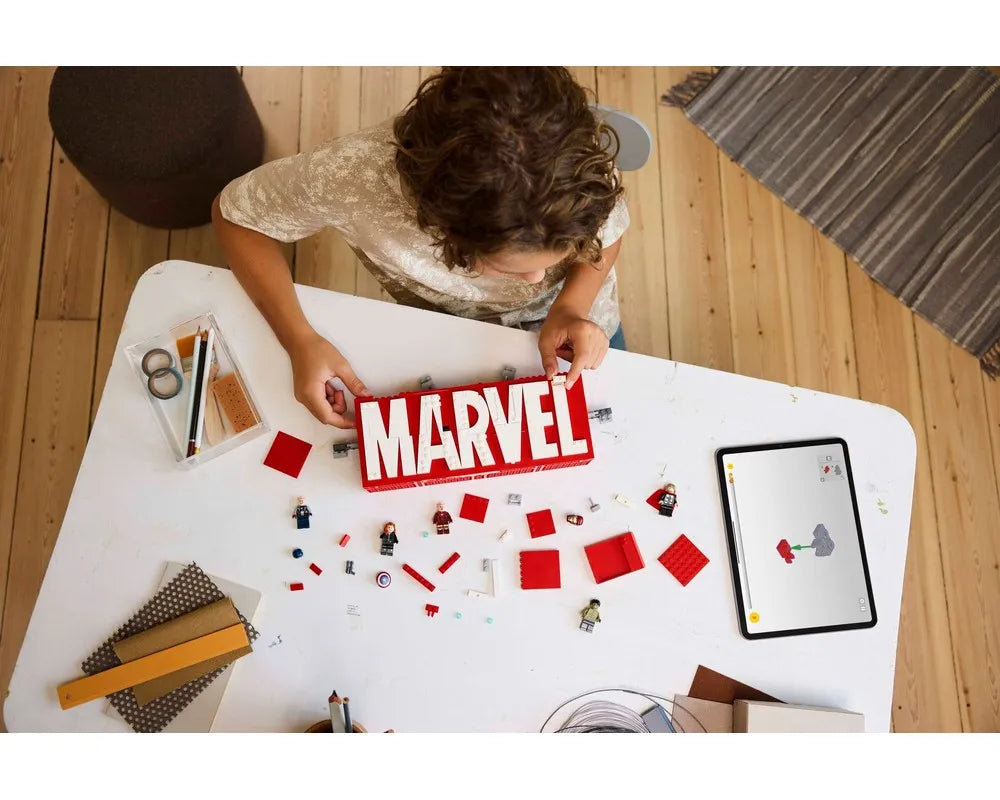 Overhead view of a child building a red and white LEGO Marvel logo. Loose bricks, minifigures, and a tablet on a white table.