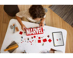 Overhead view of a child building a red and white LEGO Marvel logo. Loose bricks, minifigures, and a tablet on a white table.